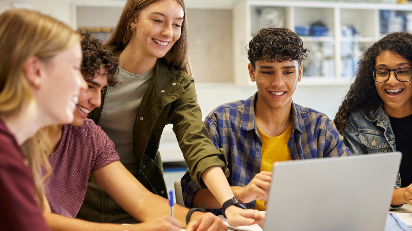 Image of a group of students gathered around a laptop in a classroom, working together on a project with notebooks and pens on the table.