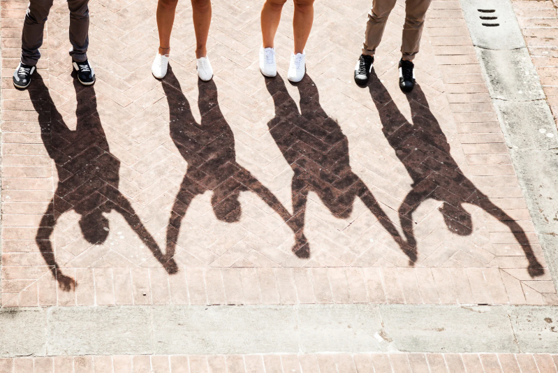 Image of Shadows of four people standing and holding hands on a sunlit brick pavement, with legs and shoes above the shadows.